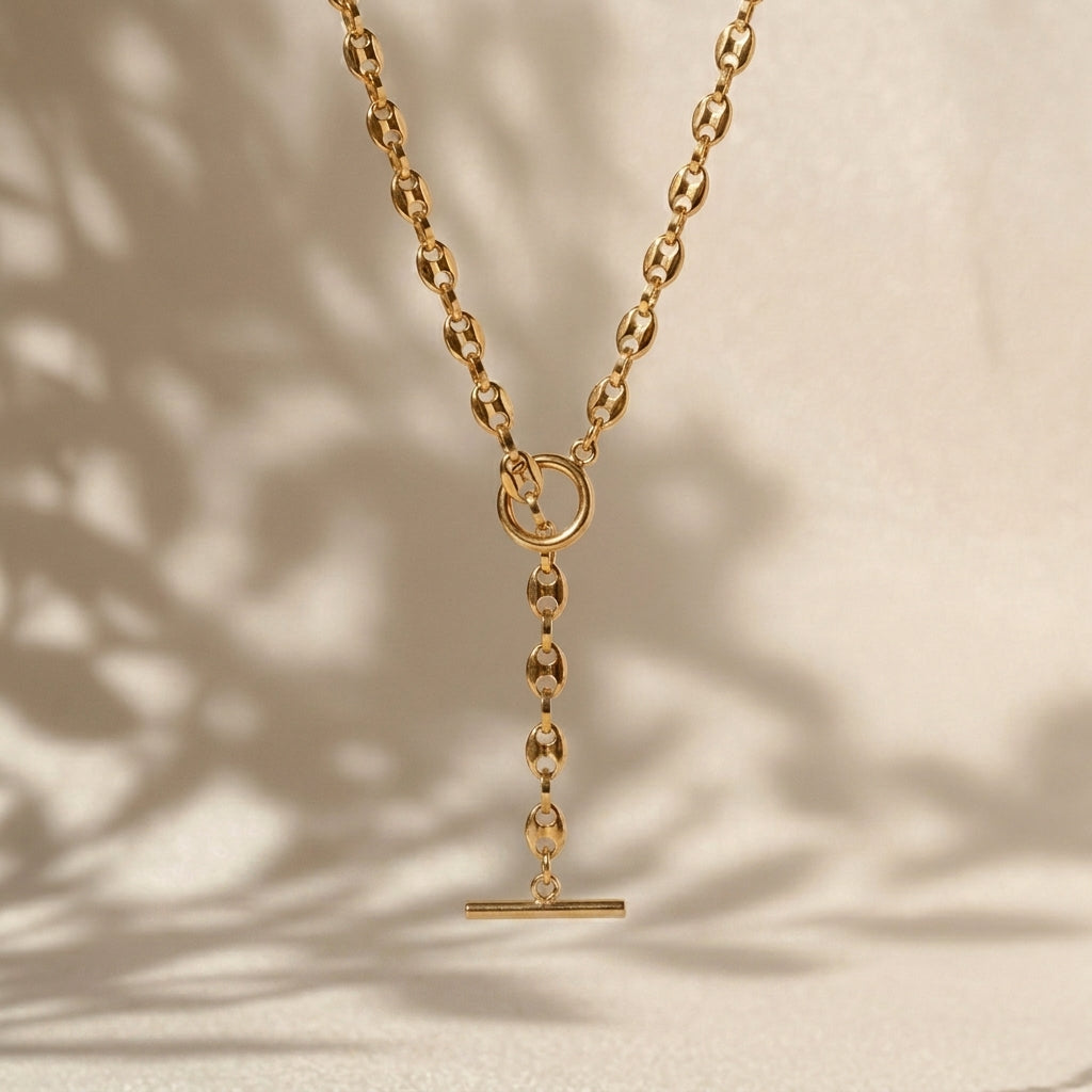 A gold-toned Y-necklace with a flat circular ring at the top, a short chain drop, and a horizontal bar pendant, displayed against a warm beige background with soft leaf shadows.