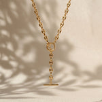 A gold-toned Y-necklace with a flat circular ring at the top, a short chain drop, and a horizontal bar pendant, displayed against a warm beige background with soft leaf shadows.