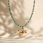 A gold crab pendant necklace with a beaded chain featuring turquoise and olive green faceted beads, displayed against a warm beige background with soft leaf shadows.