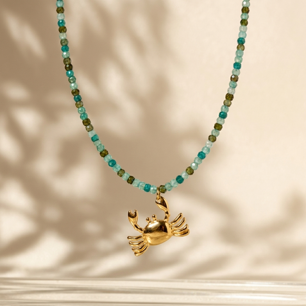 A gold crab pendant necklace with a beaded chain featuring turquoise and olive green faceted beads, displayed against a warm beige background with soft leaf shadows.