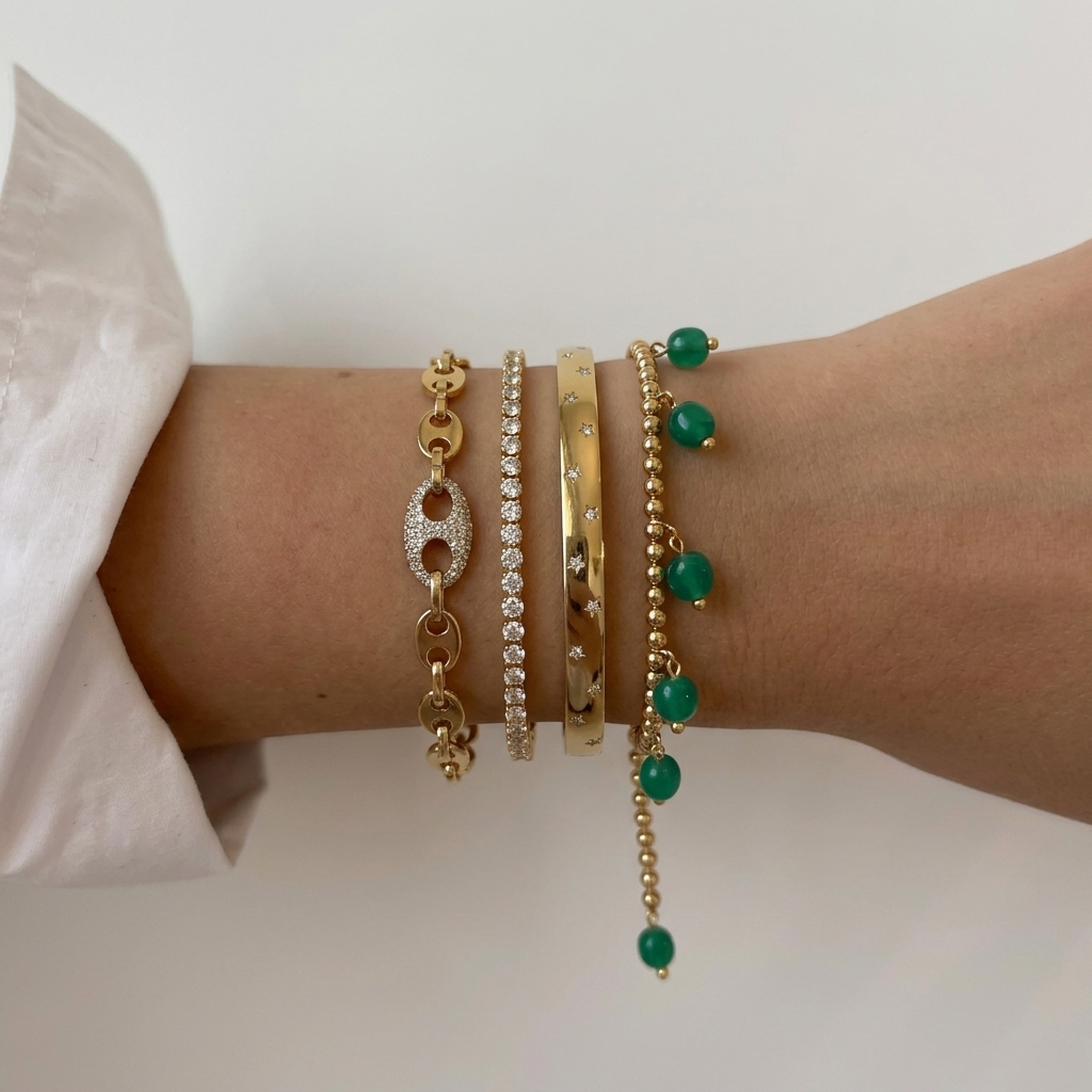 A close-up of a person's wrist wearing four delicate gold bracelets: a link bracelet with a clear crystal H charm, a pavé crystal bracelet, a smooth gold bangle, and a gold beaded bracelet with green bead drops.