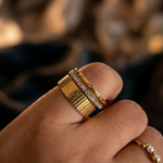 A close-up of a hand wearing a stack of gold rings, including a wide ribbed band, a slim diamond-accented band, and a gold band with a small pearl detail, against a warm, blurred background.