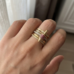 A close-up of a hand wearing a stack of gold rings, including one with a central round pink gemstone and others with small clear stones, set against a softly blurred indoor background.