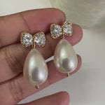 Gold CZ bow stud earrings with large teardrop baroque pearl drop held in hand against white background