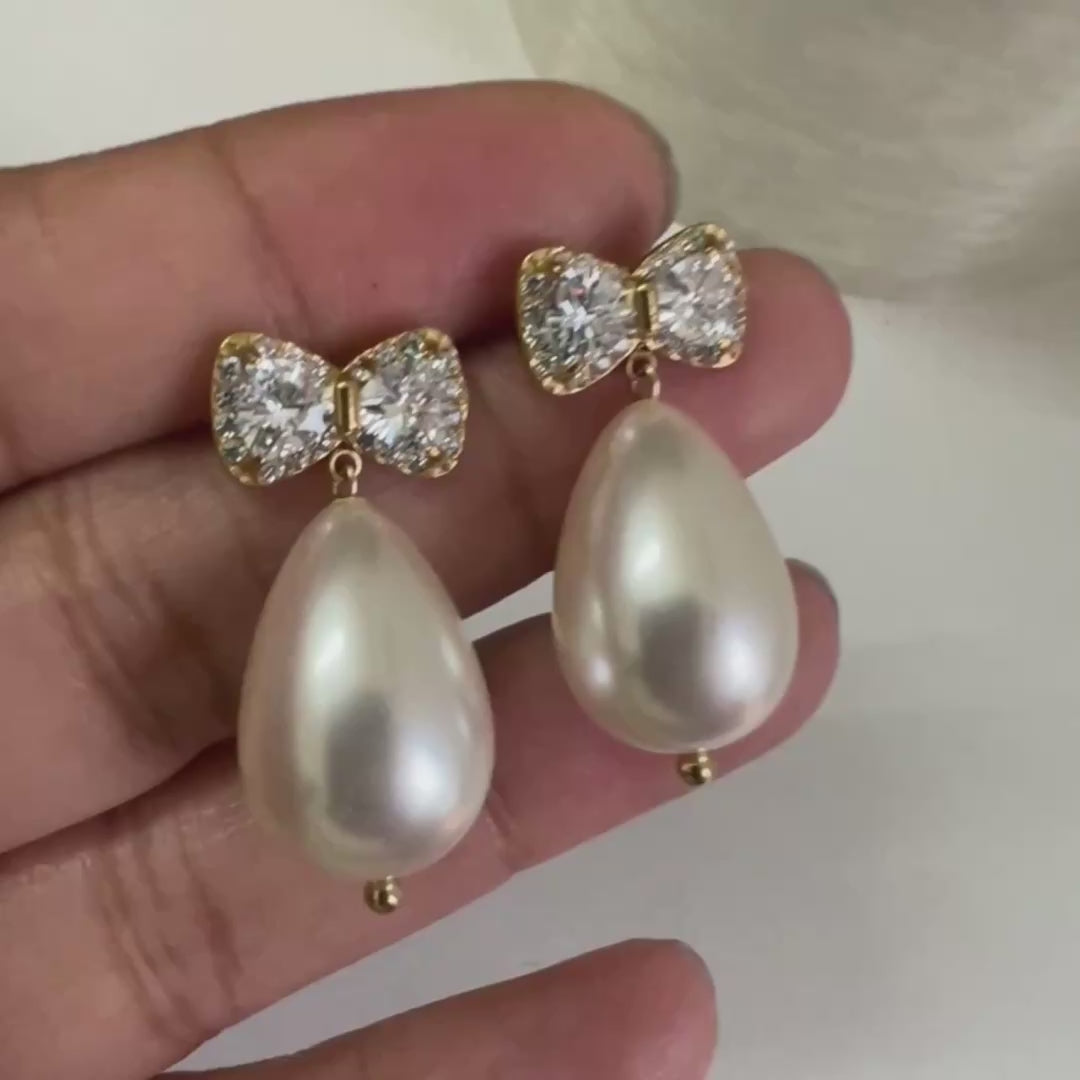Gold CZ bow stud earrings with large teardrop baroque pearl drop held in hand against white background