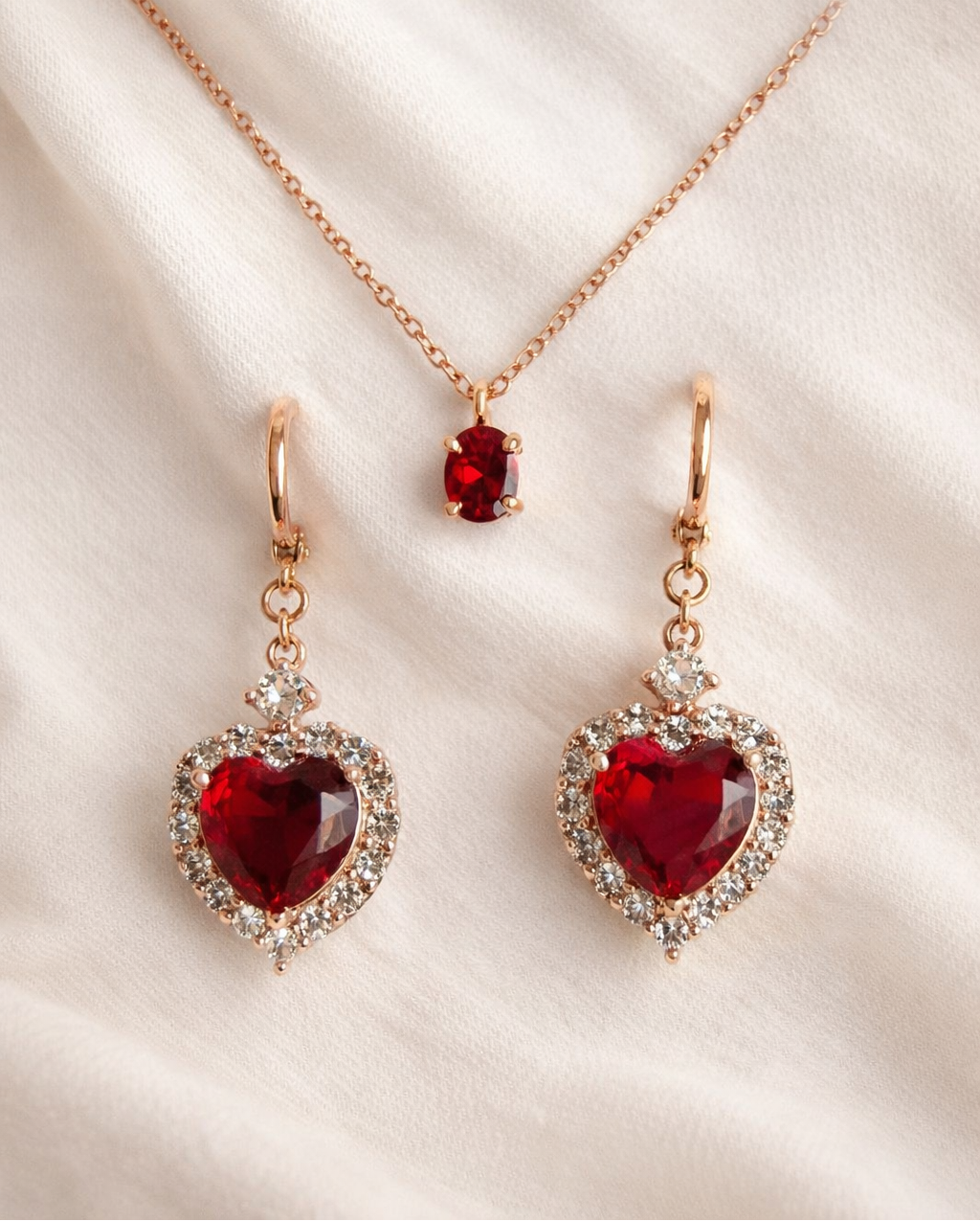 Rose gold jewelry set with red heart-shaped gemstones on a beige fabric background