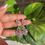 Pink and silver earrings held by a hand with green foliage in the background