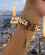 Gold bracelet with a small Eiffel Tower charm worn on a wrist, with a blurred cityscape background.